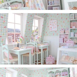 K&ouml;nnte beinhalten: Eine Miniatur-B&auml;ckerei-Szene mit einer Puppe in einem Blumenkleid. Der Raum verf&uuml;gt &uuml;ber eine pastellgr&uuml;ne und rosa Einrichtung, darunter karierte Tapeten, Vorh&auml;nge und M&ouml;bel. Regale zeigen Schachteln und Backwaren, mit einer Vitrine voller Geb&auml;ck.