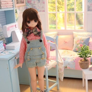K&ouml;nnte beinhalten: Eine Puppe in einem Miniaturzimmer, bekleidet mit einem rosa Polka-Dot-Hemd, Jeans-Latzhosen und Turnschuhen. Das Zimmer verf&uuml;gt &uuml;ber einen blauen Schreibtisch, ein Fenster und ein kleines Sofa mit floralen Kissen. Die Puppe hat braunes Haar und eine karierte Fliege.