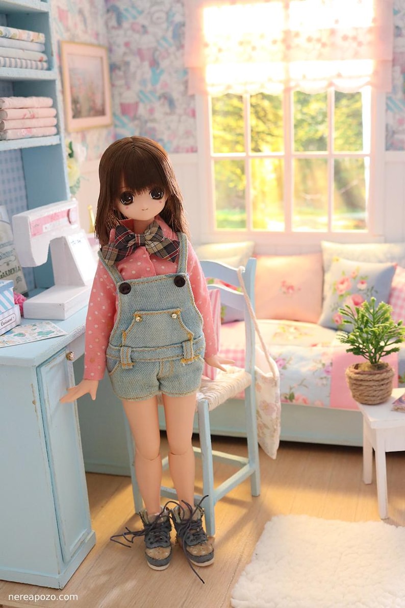 K&ouml;nnte beinhalten: Eine Puppe in einem Miniaturzimmer, bekleidet mit einem rosa Polka-Dot-Hemd, Jeans-Latzhosen und Turnschuhen. Das Zimmer verf&uuml;gt &uuml;ber einen blauen Schreibtisch, ein Fenster und ein kleines Sofa mit floralen Kissen. Die Puppe hat braunes Haar und eine karierte Fliege.