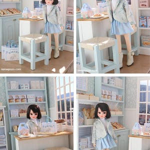 May include: A doll with dark hair wearing a light blue patterned jacket, white turtleneck, and light blue skirt, standing in a miniature bakery scene with light blue shelves, a wooden counter, woven stools, and small bakery-themed boxes and bags.