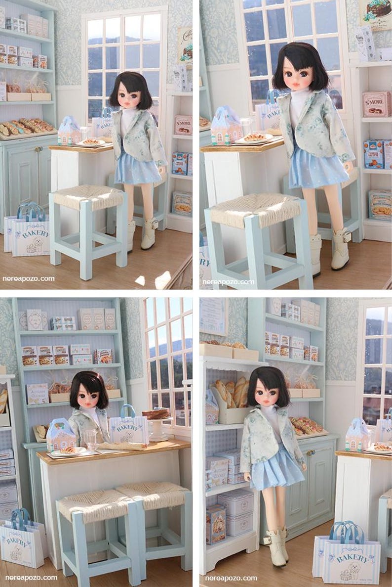 May include: A doll with dark hair wearing a light blue patterned jacket, white turtleneck, and light blue skirt, standing in a miniature bakery scene with light blue shelves, a wooden counter, woven stools, and small bakery-themed boxes and bags.