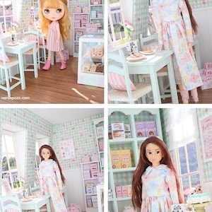 K&ouml;nnte beinhalten: Eine Miniatur-B&auml;ckerei-Szene mit zwei Puppen. Der Raum hat hellblaue M&ouml;bel, rosa und wei&szlig; karierte Tapeten und Regale mit Miniatur-Backwaren. Eine Puppe tr&auml;gt ein Blumenkleid, die andere ein rosa gestreiftes Kleid.