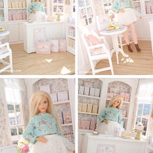 K&ouml;nnte beinhalten: Eine Miniatur-Puppenhaus-B&auml;ckerei mit einer blonden Puppe in einem lang&auml;rmeligen Blumenoberteil und einem wei&szlig;en Spitzenrock. Der Laden enth&auml;lt wei&szlig;e Regale, eine Verkaufstheke, kleine Tische, St&uuml;hle und Einkaufst&uuml;ten mit der Aufschrift Cake & Clover.