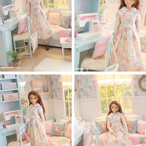 K&ouml;nnte beinhalten: Eine Puppe in einem Blumenkleid steht in einem Miniaturzimmer mit einer N&auml;hmaschine, einem Stuhl und einem Sofa. Der Raum ist in Pastellfarben mit Blumentapeten und gerahmten Bildern dekoriert.