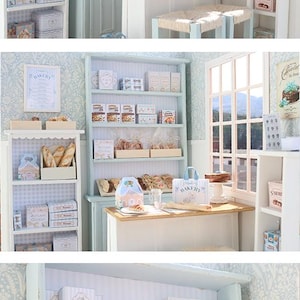 May include: A miniature bakery diorama featuring light blue wooden shelving units filled with small boxes of cookies, bread loaves, and pastries. The scene includes a small wooden table with stools, a window, and various printed signs labeled Bakery, S'more, and Farmhouse.