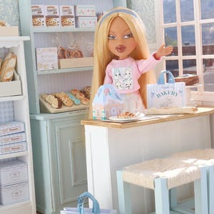 May include: A blonde doll with a blue headband wearing a pink long-sleeve shirt stands behind a white counter in a miniature bakery. The scene includes light blue shelving units, assorted miniature baked goods, and paper bags labeled Bakery.