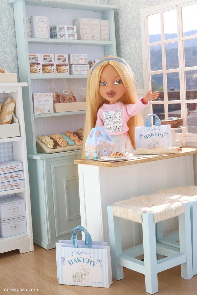 May include: A blonde doll with a blue headband wearing a pink long-sleeve shirt stands behind a white counter in a miniature bakery. The scene includes light blue shelving units, assorted miniature baked goods, and paper bags labeled Bakery.