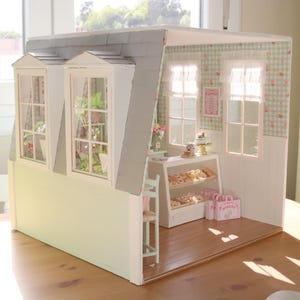 K&ouml;nnte beinhalten: Ein Miniatur-Puppenhaus-B&auml;ckerei mit grauem Dach, wei&szlig;en W&auml;nden und drei Dachgauben. Im Inneren befindet sich eine Vitrine mit Geb&auml;ck, und rosa Einkaufstaschen stehen auf dem Boden. Der Innenraum ist mit floralen Tapeten und Vorh&auml;ngen dekoriert.