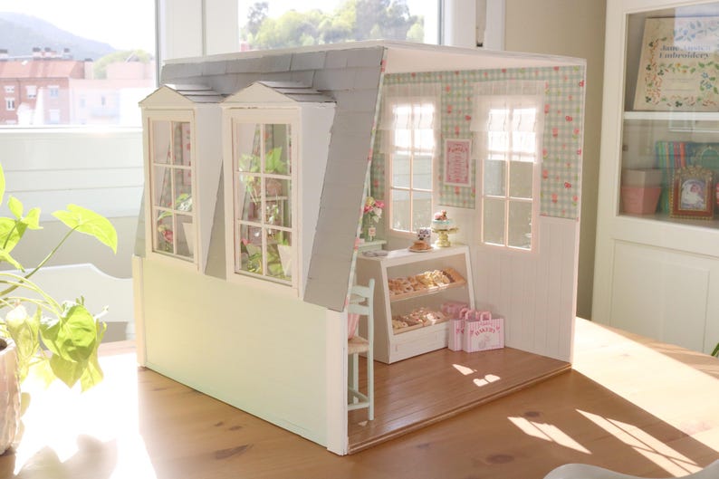 K&ouml;nnte beinhalten: Ein Miniatur-Puppenhaus-B&auml;ckerei mit grauem Dach, wei&szlig;en W&auml;nden und drei Dachgauben. Im Inneren befindet sich eine Vitrine mit Geb&auml;ck, und rosa Einkaufstaschen stehen auf dem Boden. Der Innenraum ist mit floralen Tapeten und Vorh&auml;ngen dekoriert.
