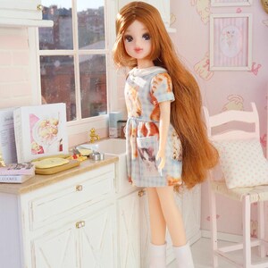 May include: A doll with long red hair wearing a blue and white patterned dress and light blue shoes. The doll is in a miniature kitchen with white cabinets and a window. The dress has a cat print.