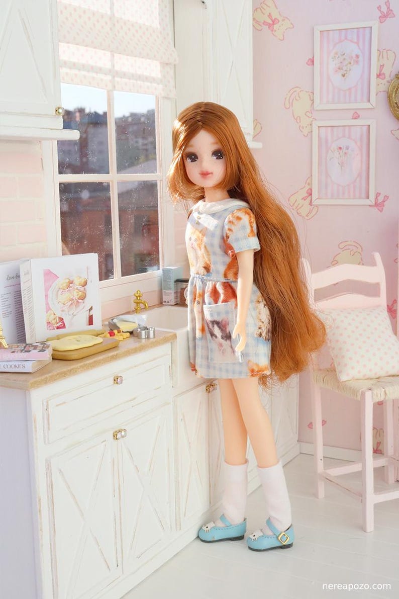 May include: A doll with long red hair wearing a blue and white patterned dress and light blue shoes. The doll is in a miniature kitchen with white cabinets and a window. The dress has a cat print.