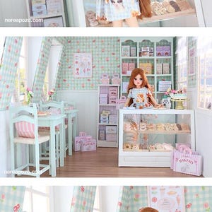 K&ouml;nnte beinhalten: Eine Puppe in einem blauen Kleid mit Katzenmuster steht in einer Miniatur-B&auml;ckerei. Die B&auml;ckerei verf&uuml;gt &uuml;ber Regale mit Schachteln, eine Vitrine mit Geb&auml;ck und Tische. Die W&auml;nde sind mit einem hellblau-wei&szlig;en Karomuster verziert.