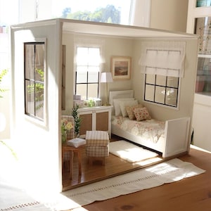 K&ouml;nnte beinhalten: Ein Miniatur-Puppenhauszimmer mit einem wei&szlig;en Bett mit gemusterter Tagesdecke, einem kleinen Holztisch, einem karierten Hocker und einem wei&szlig;en Schrank. Der Raum verf&uuml;gt &uuml;ber zwei Fenster mit schwarzen Rahmen, wei&szlig;en Vorh&auml;ngen und einer Lampe.