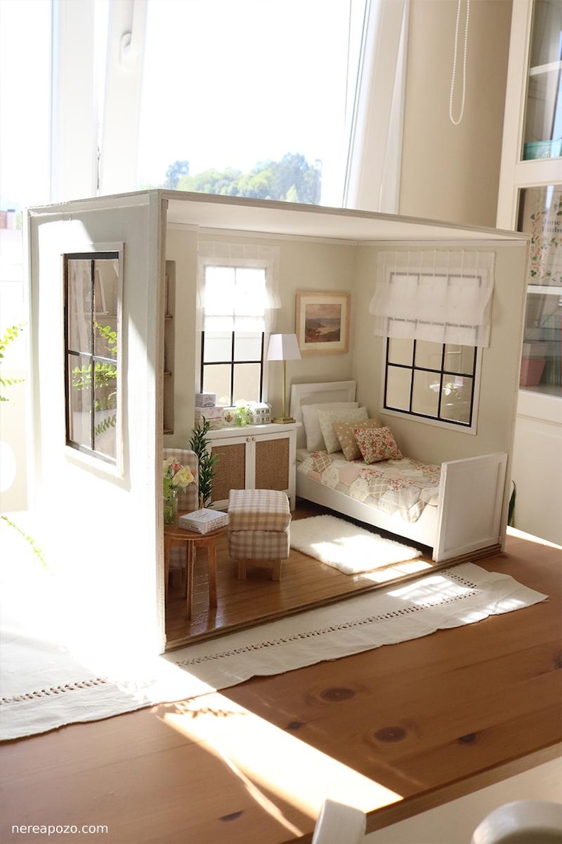 K&ouml;nnte beinhalten: Ein Miniatur-Puppenhauszimmer mit einem wei&szlig;en Bett mit gemusterter Tagesdecke, einem kleinen Holztisch, einem karierten Hocker und einem wei&szlig;en Schrank. Der Raum verf&uuml;gt &uuml;ber zwei Fenster mit schwarzen Rahmen, wei&szlig;en Vorh&auml;ngen und einer Lampe.