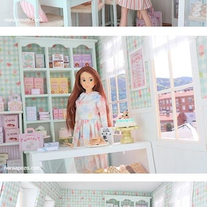 K&ouml;nnte beinhalten: Eine Miniatur-B&auml;ckerei-Szene mit Puppen, in einem hellblauen und rosa Farbschema. Die Szene umfasst einen Tisch und St&uuml;hle, Regale mit Miniatur-Backwaren und eine Vitrine. Die W&auml;nde sind mit einem floralen Muster verziert.