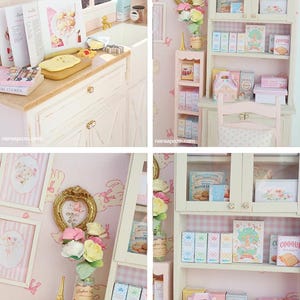 May include: A miniature bakery scene with a white cabinet, shelves, and a small pink chair. The shelves are filled with boxes, books, and decorative items. The walls are pink with framed pictures and a floral arrangement. A small kitchen area is also visible.