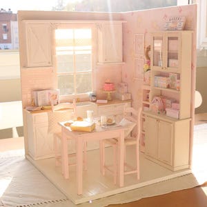 May include: A miniature kitchen in pink and white, featuring a table, chairs, and cabinets. A small cake is on the counter. The word "BAKERY" is on a sign. The scene is brightly lit, creating a cheerful and welcoming feel.