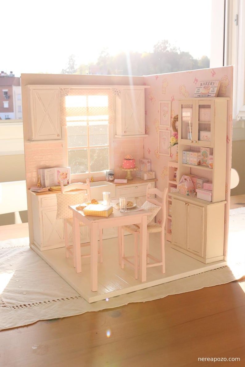 May include: A miniature kitchen in pink and white, featuring a table, chairs, and cabinets. A small cake is on the counter. The word "BAKERY" is on a sign. The scene is brightly lit, creating a cheerful and welcoming feel.