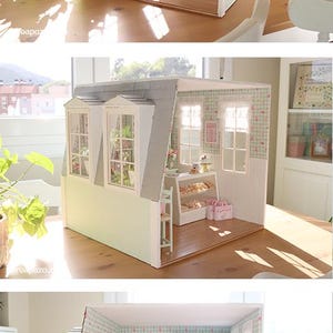 K&ouml;nnte beinhalten: Miniatur-B&auml;ckerei-Szene in Hellgr&uuml;n und Rosa. Das Puppenhaus zeigt eine Vitrine mit Geb&auml;ck, Tischen, St&uuml;hlen und Regalen voller Schachteln. Die W&auml;nde sind mit einem floralen Muster verziert.