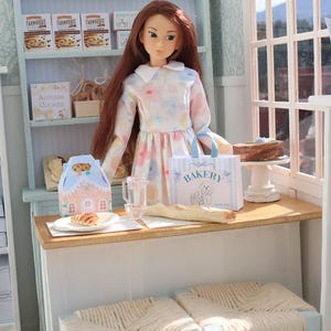 May include: A doll with long brown hair wearing a floral dress stands behind a counter in a miniature bakery. The counter holds a small bakery bag with the text BAKERY, a glass pitcher, a baguette, and a pastry. Shelves display cookie boxes.