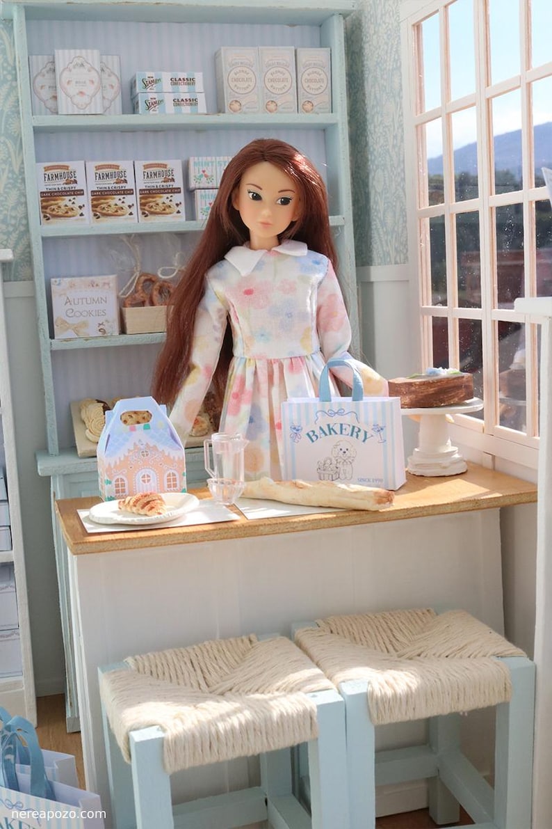 May include: A doll with long brown hair wearing a floral dress stands behind a counter in a miniature bakery. The counter holds a small bakery bag with the text BAKERY, a glass pitcher, a baguette, and a pastry. Shelves display cookie boxes.