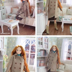 Może przedstawiać: Lalka z długimi rudymi włosami w beżowym trenczu, ciemnej sp&oacute;dnicy i beżowych butach. Lalka znajduje się w miniaturowym pokoju z białym stołem, sofą i oknem.