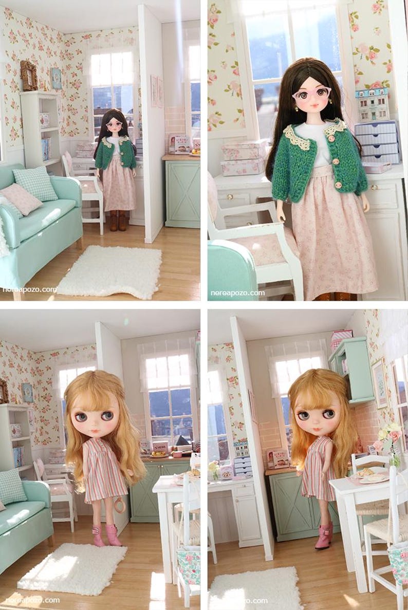 May include: A dollhouse scene with a doll in a pink dress and green jacket. The doll is in a miniature room with floral wallpaper, a sofa, and a small table. The room is decorated in pastel colours.