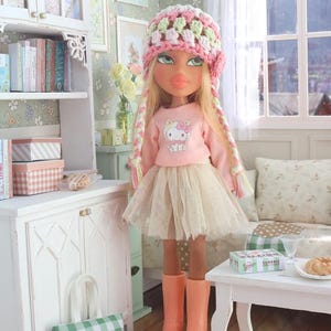 Może przedstawiać: Lalka Bratz w r&oacute;żowym swetrze Hello Kitty, beżowej tiulowej sp&oacute;dnicy i r&oacute;żowych butach, z r&oacute;żową, zieloną i białą czapką z szydełka i warkoczami. Lalka znajduje się w miniaturowym pokoju z białą szafką i małym stolikiem.