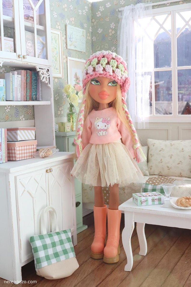 Może przedstawiać: Lalka Bratz w r&oacute;żowym swetrze Hello Kitty, beżowej tiulowej sp&oacute;dnicy i r&oacute;żowych butach, z r&oacute;żową, zieloną i białą czapką z szydełka i warkoczami. Lalka znajduje się w miniaturowym pokoju z białą szafką i małym stolikiem.
