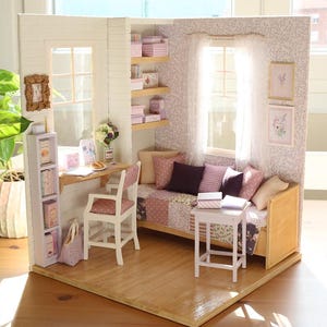 Pode incluir: Interior de casa de bonecas em miniatura com secret&aacute;ria, cadeira e sof&aacute;. O quarto apresenta uma janela, prateleiras com caixas e obras de arte emolduradas. O esquema de cores inclui rosa, branco e tons de madeira.