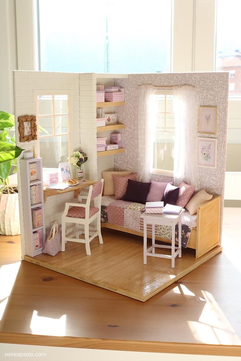 Pode incluir: Interior de casa de bonecas em miniatura com secret&aacute;ria, cadeira e sof&aacute;. O quarto apresenta uma janela, prateleiras com caixas e obras de arte emolduradas. O esquema de cores inclui rosa, branco e tons de madeira.