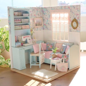 K&ouml;nnte beinhalten: Miniatur-Puppenhaus-Zimmer mit hellblauem Schreibtisch, B&uuml;cherregal und Sofa. Der Raum ist mit floraler Tapete, rosa und blauen Kissen und einer kleinen N&auml;hmaschine dekoriert. Eine Miniatur-Einkaufstasche und ein Teppich vervollst&auml;ndigen die Szene.