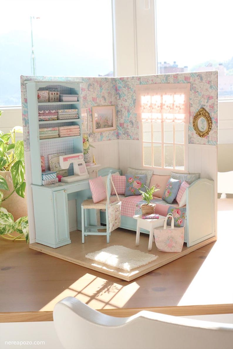 K&ouml;nnte beinhalten: Miniatur-Puppenhaus-Zimmer mit hellblauem Schreibtisch, B&uuml;cherregal und Sofa. Der Raum ist mit floraler Tapete, rosa und blauen Kissen und einer kleinen N&auml;hmaschine dekoriert. Eine Miniatur-Einkaufstasche und ein Teppich vervollst&auml;ndigen die Szene.