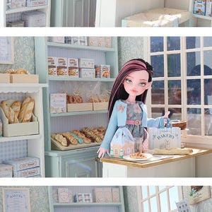 May include: A miniature bakery scene featuring a doll with long dark hair and pink highlights, wearing a light blue cardigan over a floral dress. The doll stands at a wooden counter with a small paper bag labeled Bakery and a pastry on a plate.