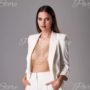 May include: A woman wearing a white blazer and white pants. She is wearing a gold chain body jewelry piece.
