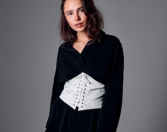 May include: A white satin corset with a lace-up front is worn over a black top. The text "Custom sizes | High-quality satin" is displayed at the bottom of the image. The corset is a waist cincher.