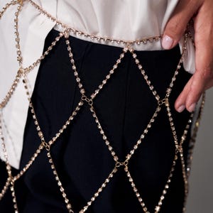 May include: Gold-toned body chain with sparkling rhinestone accents. The chain is designed in a diamond pattern, creating a decorative overlay. The body chain is worn over black pants and a white shirt.