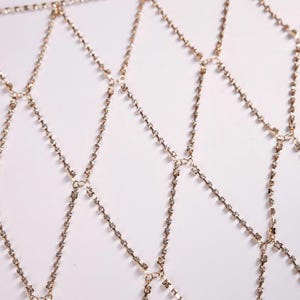May include: A gold-colored body chain with a diamond-shaped pattern. The chain is made of small, sparkling rhinestones set in a delicate, interconnected design. The piece is designed to be worn over clothing.