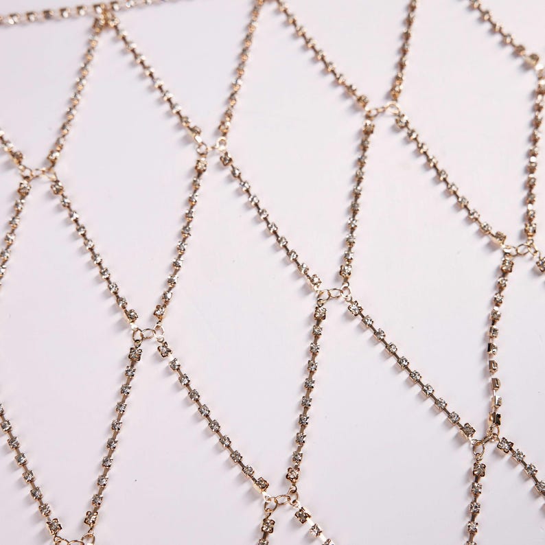 May include: A gold-colored body chain with a diamond-shaped pattern. The chain is made of small, sparkling rhinestones set in a delicate, interconnected design. The piece is designed to be worn over clothing.