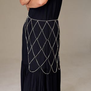 May include: A silver-colored body chain with a diamond-shaped pattern. The chain is worn over a black dress. The chain is made of small, reflective metal pieces, creating a shimmering effect. The dress is a solid black color.