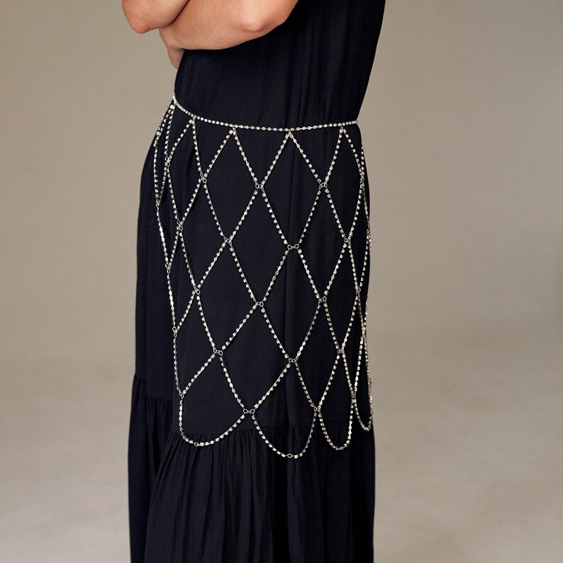 May include: A silver-colored body chain with a diamond-shaped pattern. The chain is worn over a black dress. The chain is made of small, reflective metal pieces, creating a shimmering effect. The dress is a solid black color.