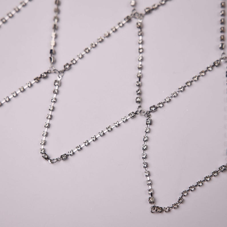 May include: Silver body jewelry featuring multiple strands of small, sparkling rhinestones. The delicate chains are connected at various points, creating a layered effect. The jewelry is designed to be worn on the body.