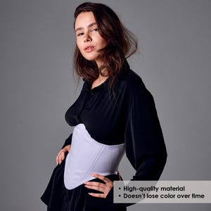 May include: A white corset with vertical seams is worn over a black long-sleeve shirt. The corset is the focal point, with a smooth, structured appearance. The image includes text that says "High-quality material" and "Doesn't lose color over time."