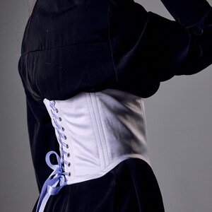 May include: A white satin corset with a light purple ribbon lace-up detail. The corset is worn over a black garment. The design features vertical stitching and a curved shape, offering a classic silhouette.