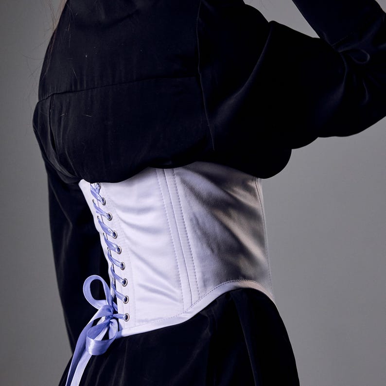 May include: A white satin corset with a light purple ribbon lace-up detail. The corset is worn over a black garment. The design features vertical stitching and a curved shape, offering a classic silhouette.