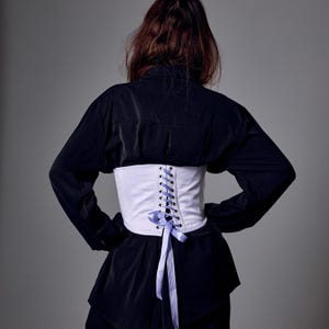 May include: A white corset with a lace-up back and a light blue ribbon. The corset is worn over a black shirt. The image is taken from the back, showing the details of the corset and the shirt.