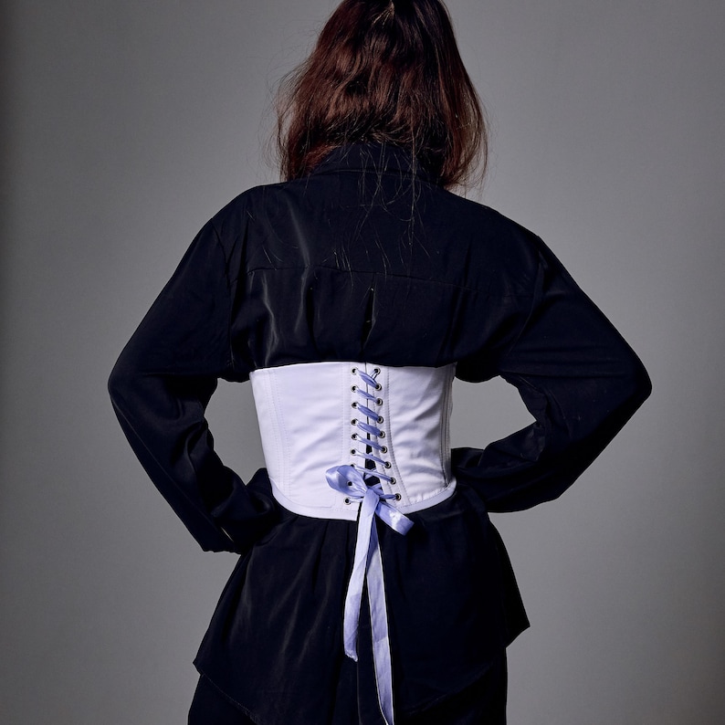 May include: A white corset with a lace-up back and a light blue ribbon. The corset is worn over a black shirt. The image is taken from the back, showing the details of the corset and the shirt.