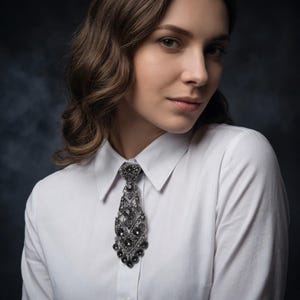 Puede incluir: Una camisa blanca de botones con un broche decorativo en forma de corbata. El broche está elaborado con filigrana de metal en tonos metálicos oscuros y adornado con numerosas piedras pequeñas facetadas de color gris oscuro en un patrón complejo.