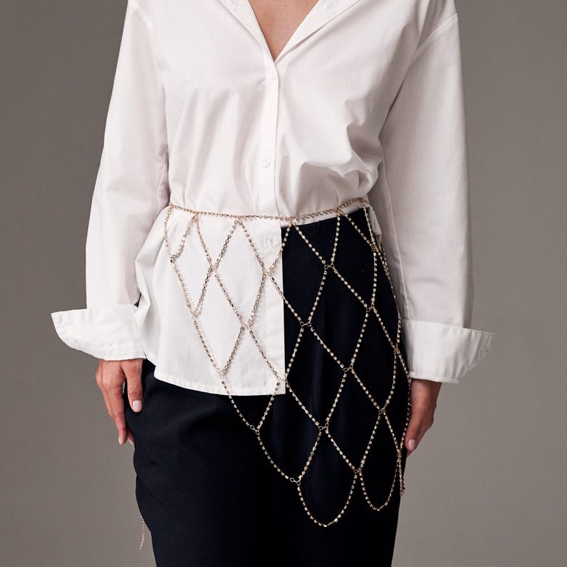 May include: A gold-colored chain belt with a diamond-shaped design. The belt is worn over a white button-down shirt and a black skirt. The chain drapes down the front of the skirt.