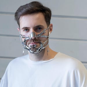 Puede incluir: Una máscara facial plateada con un diseño intrincado y calado. La máscara cubre la nariz y la parte inferior de la cara, con elementos decorativos que se extienden hasta las mejillas y la barbilla. Pequeñas cadenas con dijes colgantes cuelgan de los lados de la máscara. La persona lleva una camiseta blanca.